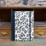Libreta. Lomo de tela negro y papel. Estampado arabesco clásico con motivos dorados y fondo beige. 